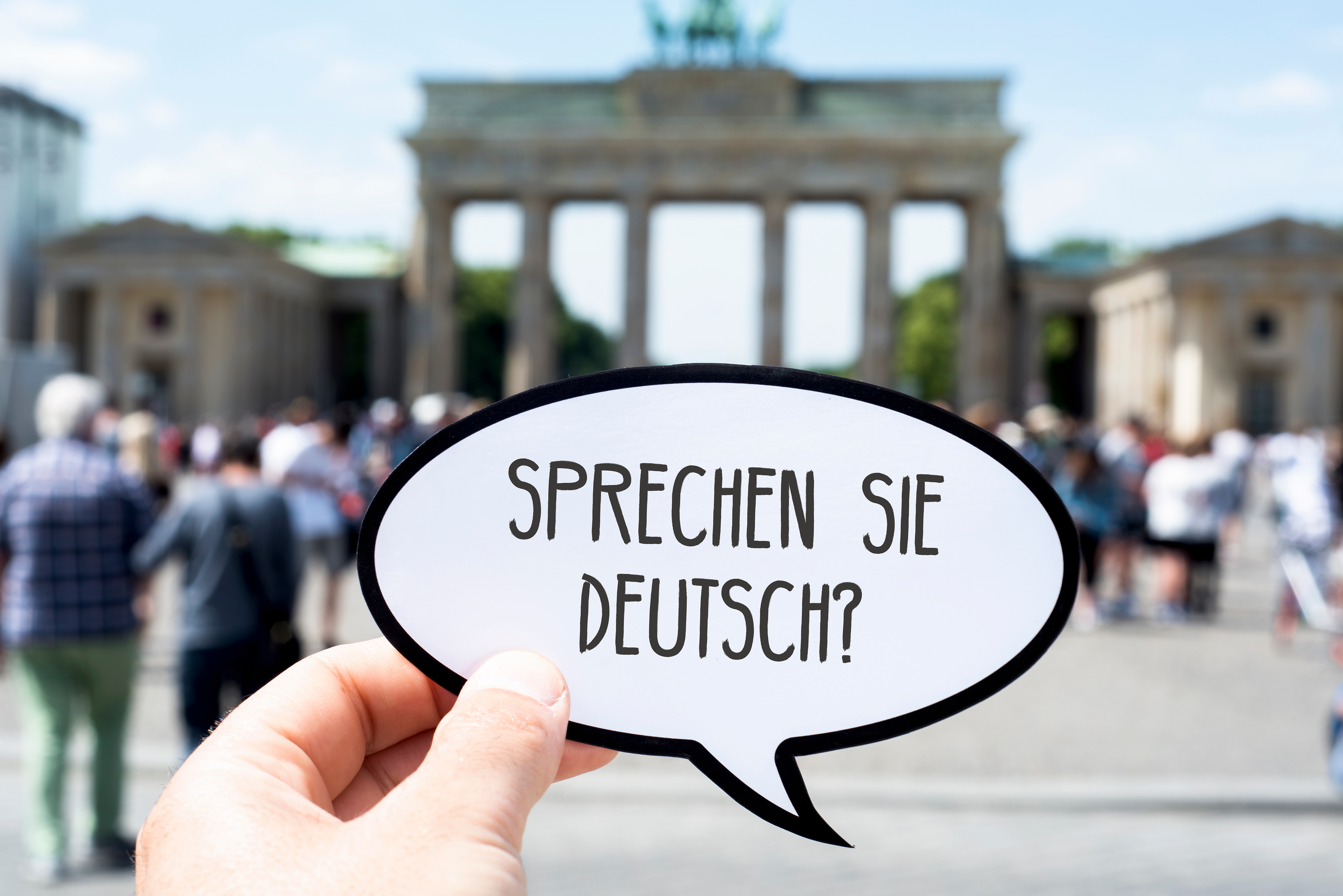 Jemand hält eine Sprechblase aus Papier mit dem Satz "Sprechen Sie Deutsch" vor das Brandenburger Tor