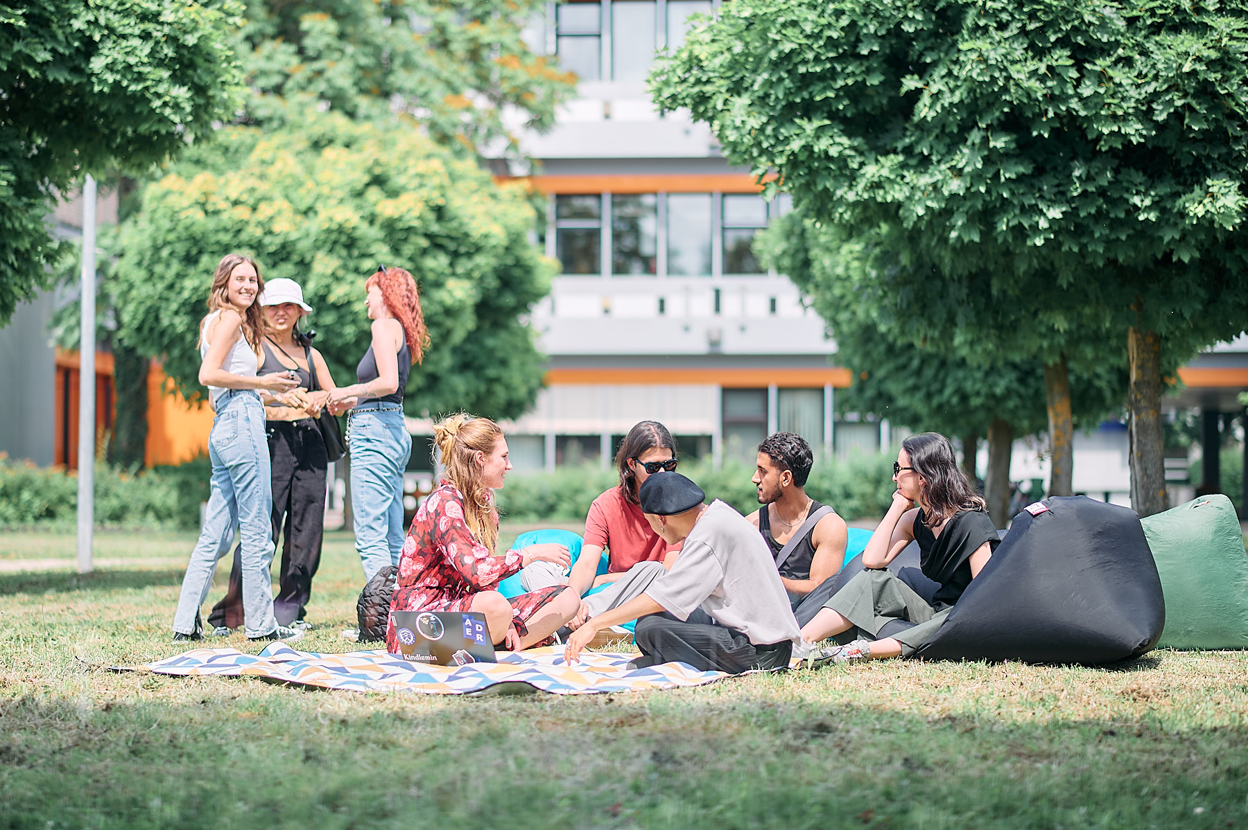 Studierende sitzen in der Sonne auf einer Wiese, unterhalten sich und spielen Ball