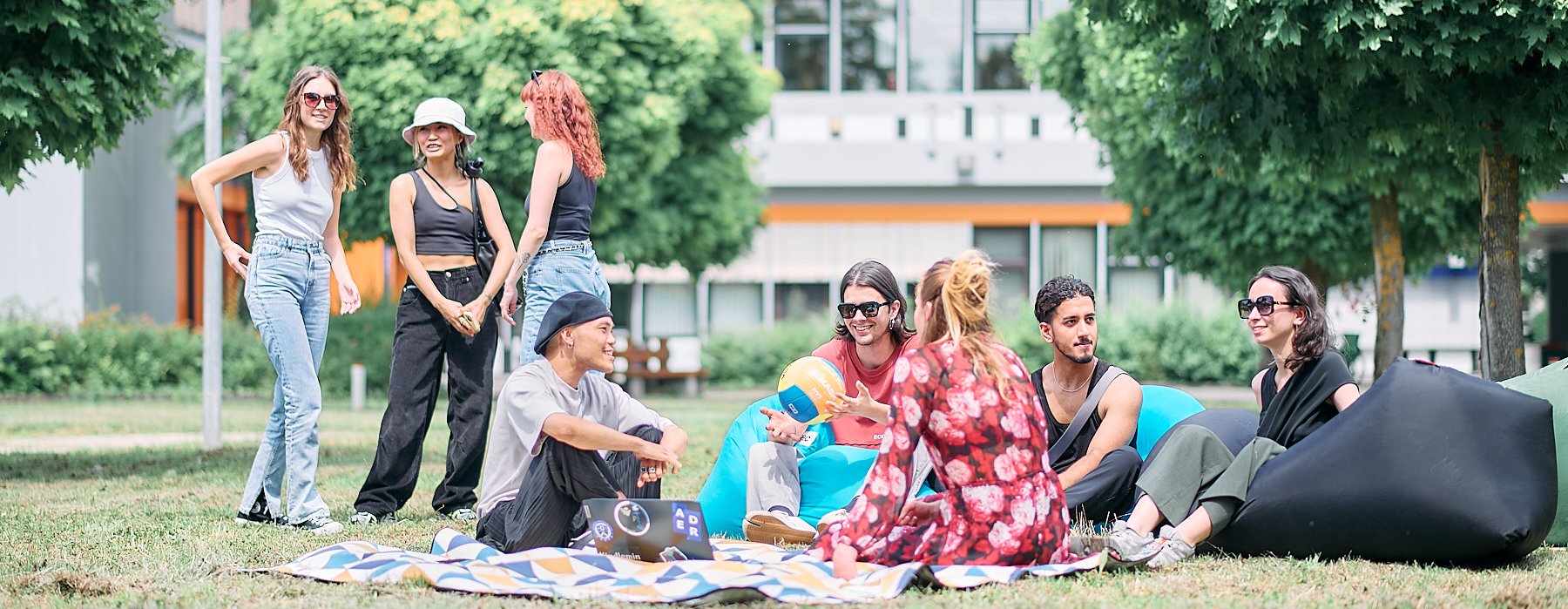 Studierende sitzen entspannt auf Decken im Gras und unterhalten sich