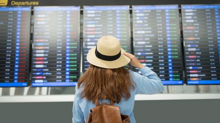 Eine Frau mit Sonnenhut und Rucksack steht vor einer digitalen Anzeige am Flughafen