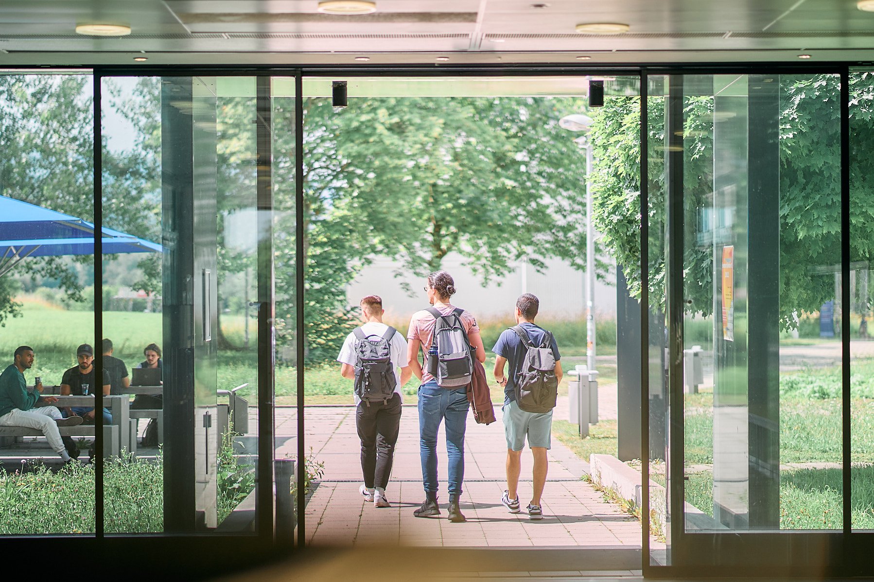 Studierende laufen von hinten gesehen aus Gebäude 3 Richtung Sitzecke mit Sonnenschirmen