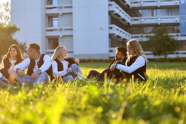 Studierende sitzen entspannt im Gras vor Gebäude 3 und genießen die Abendsonne.