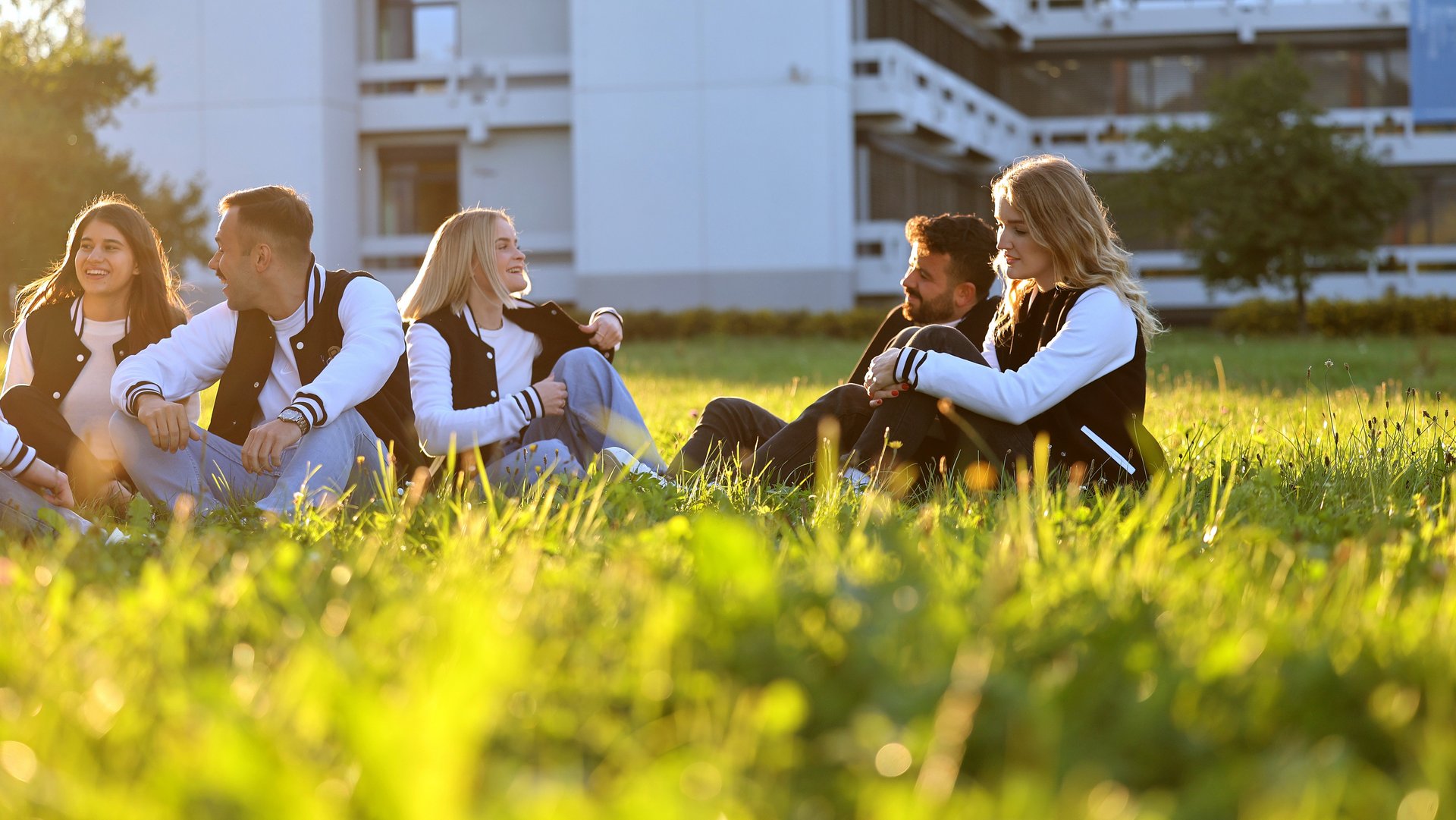 Studierende sitzen entspannt im Gras vor Gebäude 3 und genießen die Abendsonne.
